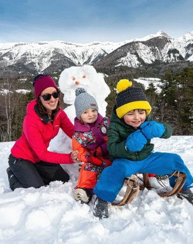 Famille sur une luge en bois devant un bonhomme de neige, avec au fond le décor de la chaîne de la Blanche