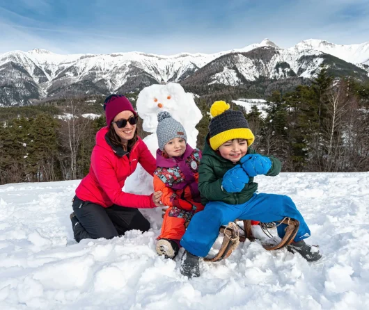 Famille sur une luge en bois devant un bonhomme de neige, avec au fond le décor de la chaîne de la Blanche