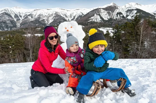 Famille sur une luge en bois devant un bonhomme de neige, avec au fond le décor de la chaîne de la Blanche